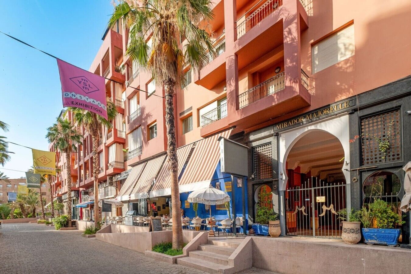 Un hotel de varios pisos con balcones, mobiliario exterior y un llamativo cartel rosa que anuncia una exposición.