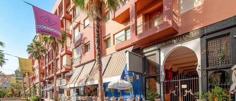 Un hotel de varios pisos con balcones, mobiliario exterior y un llamativo cartel rosa que anuncia una exposición.