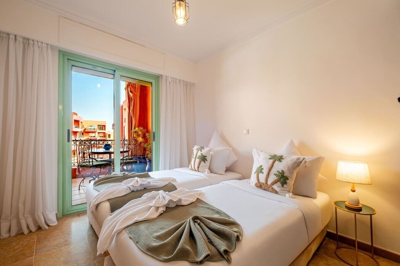 A hotel room with two beds, a balcony with a view, a table, and a lamp.
