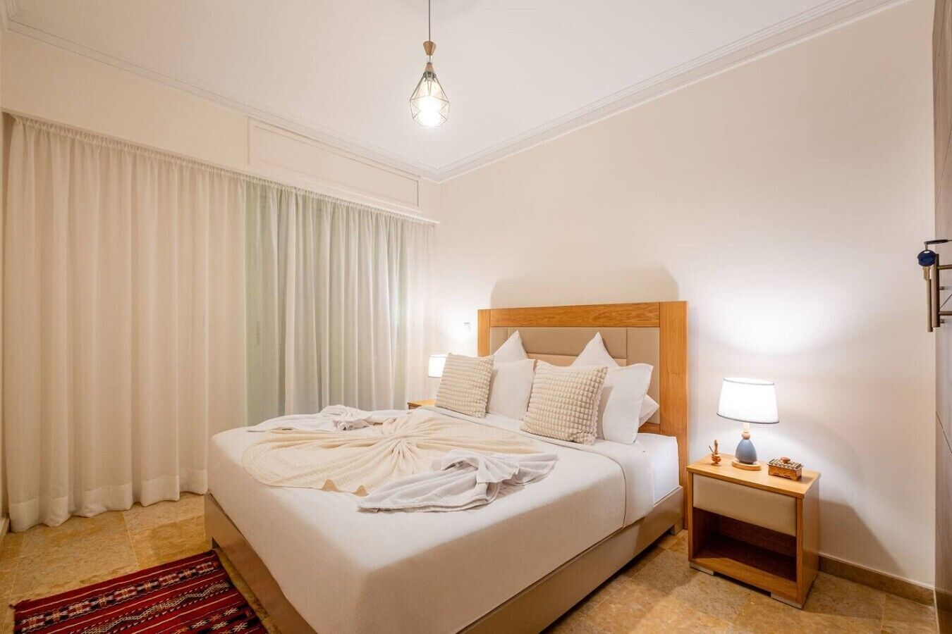 Ein Hotelzimmer mit einem großen Bett, einem Nachttisch mit Lampe und Blick ins Freie durch das Fenster.