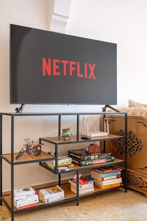 A living room with a television on a stand displaying the Netflix logo, a bookshelf with various items, and a patterned sofa. - Superbe face Jardin Majorelle | Piscine & Parking (Marrakesh)