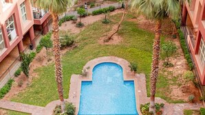 Ein rechteckiger Swimmingpool mit Steinumrandung, umgeben von einer Rasenfläche mit Palmen, neben einem Gebäude mit Balkonen.