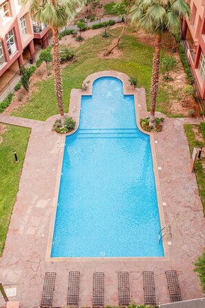 A rectangular swimming pool with a stone border, surrounded by a grassy area with palm trees, and adjacent to a building with balconies. - Superbe face Jardin Majorelle | Piscine & Parking (Marrakesh)