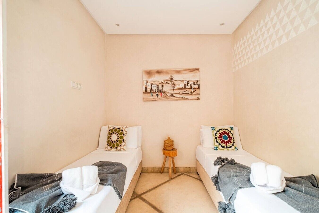 A small, single-room hotel room with two beds, a small wooden stool, and a wall-mounted painting.