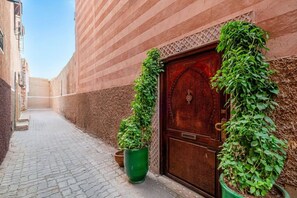 A narrow alleyway with a cobblestone path, a wooden door with intricate carvings, and two potted plants on either side. - Riad Jad • 5BR • Pool • Breakfast • Rooftop (Marrakesh)