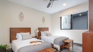 Quarto de hotel com duas camas, um ventilador de teto, uma janela e uma pequena mesa de madeira.
