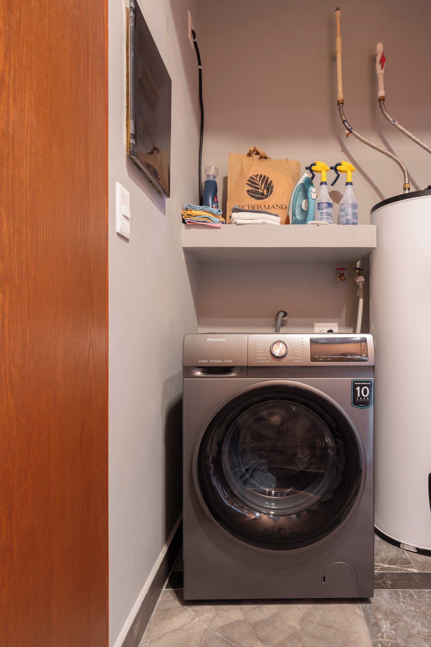 A compact laundry area with a front-loading washing machine, a shelf with cleaning supplies, and a wooden door.