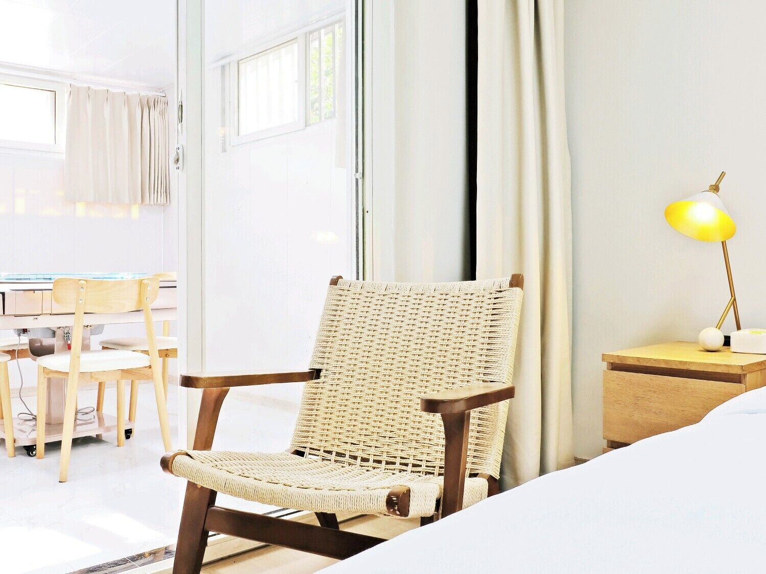 Quarto de hotel com uma cama, uma cadeira de vime, uma escrivaninha com cadeira, uma mesa de cabeceira com abajur e uma janela com cortinas.