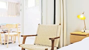 Quarto de hotel com uma cama, uma cadeira de vime, uma escrivaninha com cadeira, uma mesa de cabeceira com abajur e uma janela com cortinas.