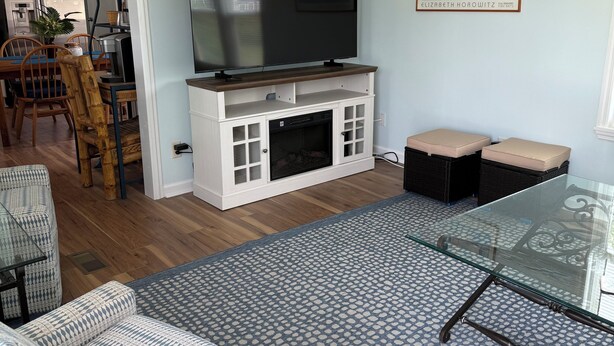 Una sala de estar con un televisor de pantalla plana sobre un mueble blanco, una mesa de centro de vidrio, sofás estampados y una zona de comedor con sillas y una mesa de madera.