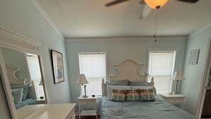 Un dormitorio con cama, cómoda, mesitas de noche, lámparas, ventilador de techo y ventana con persianas.