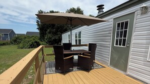 Una terraza de madera con una mesa y sillas, una pérgola con toldo, y una casa al fondo.