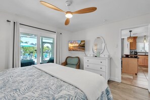 Un dormitorio con cama, ventilador de techo, cómoda, silla, un cuadro y vistas al exterior.
