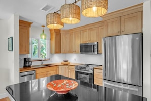 A modern kitchen with wooden cabinets, stainless steel appliances, a black countertop, and a central island with a decorative bowl. - Sunrise Lagoon by Duncan Real Estate (Anna Maria)