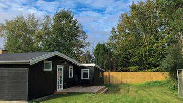 Une petite cabane de couleur sombre, dotée d’un porche et entourée d’une clôture en bois, se trouve dans une cour verdoyante.