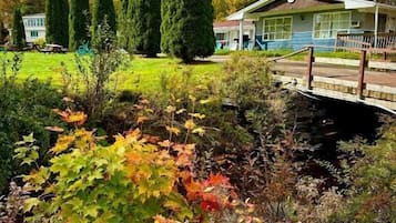 Une maison avec un porche à l’avant, un petit jardin avec différentes plantes et un pont menant à la maison.
