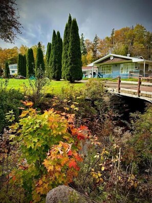 A house with a front porch, a small garden with various plants, and a bridge leading to the house. - Aberdeen Motel (Whycocomagh)
