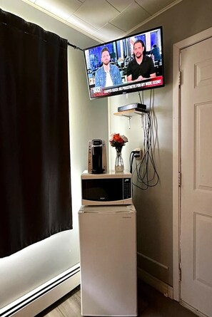A room with a television mounted on the wall displaying a news program, a small white cabinet with a flower vase, and a door to the right.