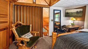 Ein gemütliches Zimmer mit Holzbalkendecke, einem Schaukelstuhl mit Kissen, einem Bett, einem Schreibtisch mit Lampe und einem Bild an der Wand.