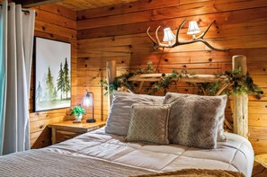 Un dormitorio de cabaña de madera con cama, almohadas, una mesita de noche con lámpara, un cuadro enmarcado y cabecera con cornamenta de ciervo.