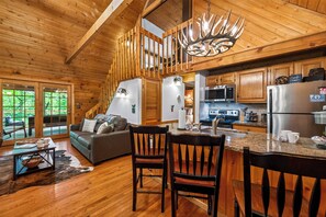 Interior de una cabaña de madera con cocina, comedor y sala de estar, que incluye un sofá y sillas.