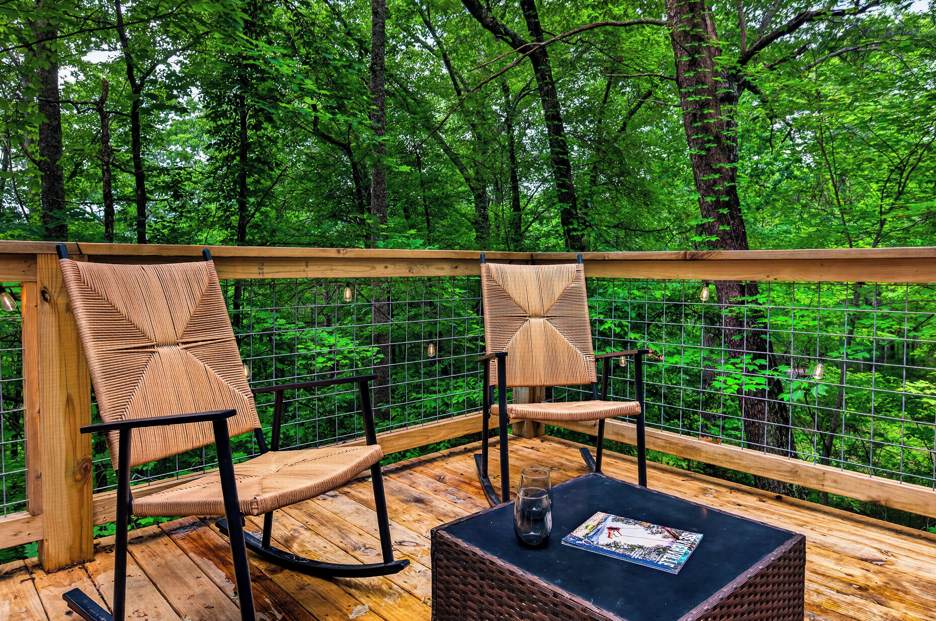 A wooden deck with two wicker chairs and a table, surrounded by a metal fence overlooking a forest.