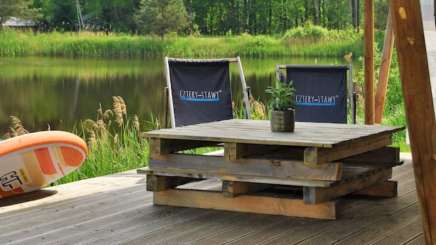 Una terraza de madera con dos sillas y una mesa, con vista a un lago tranquilo y un bosque al fondo.