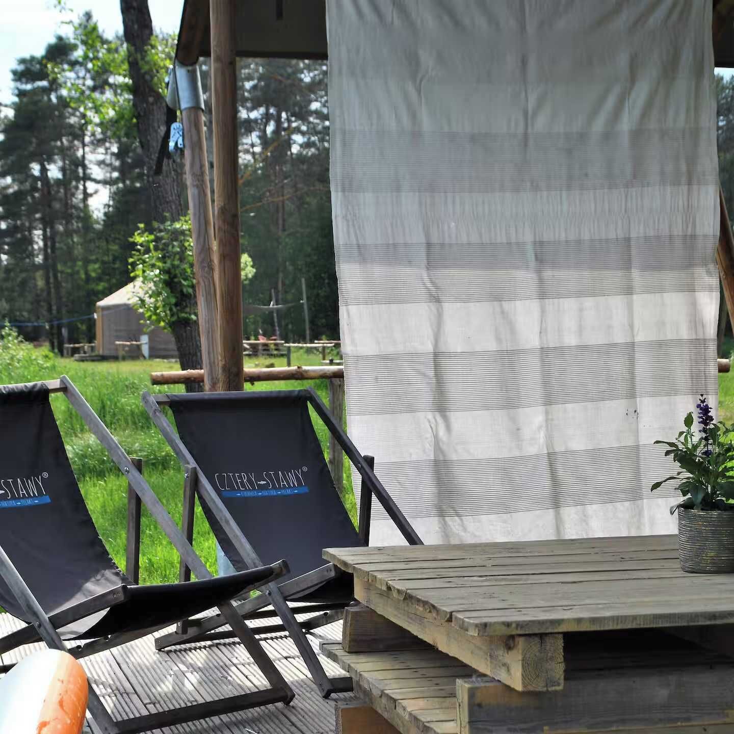 A wooden deck with two lounge chairs and a table, surrounded by a white curtain and a potted plant.