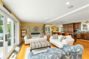A spacious living area with a stone fireplace, white sofas, and a kitchen with wooden cabinets. - NEW FOR 2027 Lake Winnipesaukee Waterfront Sleeps 12 (Moultonborough)