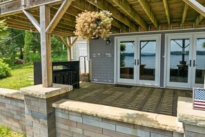 Eine Terrasse im Freien mit Grill, einer hängenden Blumenampel und Blick auf einen See.