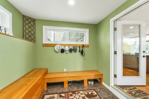 Ein Zimmer mit einer Holzbank, einem Schuhregal mit mehreren Schuhpaaren, einem Fenster mit Jalousien und einer Tür zu einem weiteren Bereich.