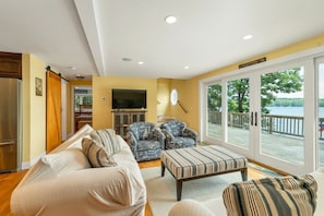 Ein Wohnzimmer mit einer Couch, zwei Sesseln, einem Fernseher und einer großen Schiebeglastür, die auf eine Terrasse führt.