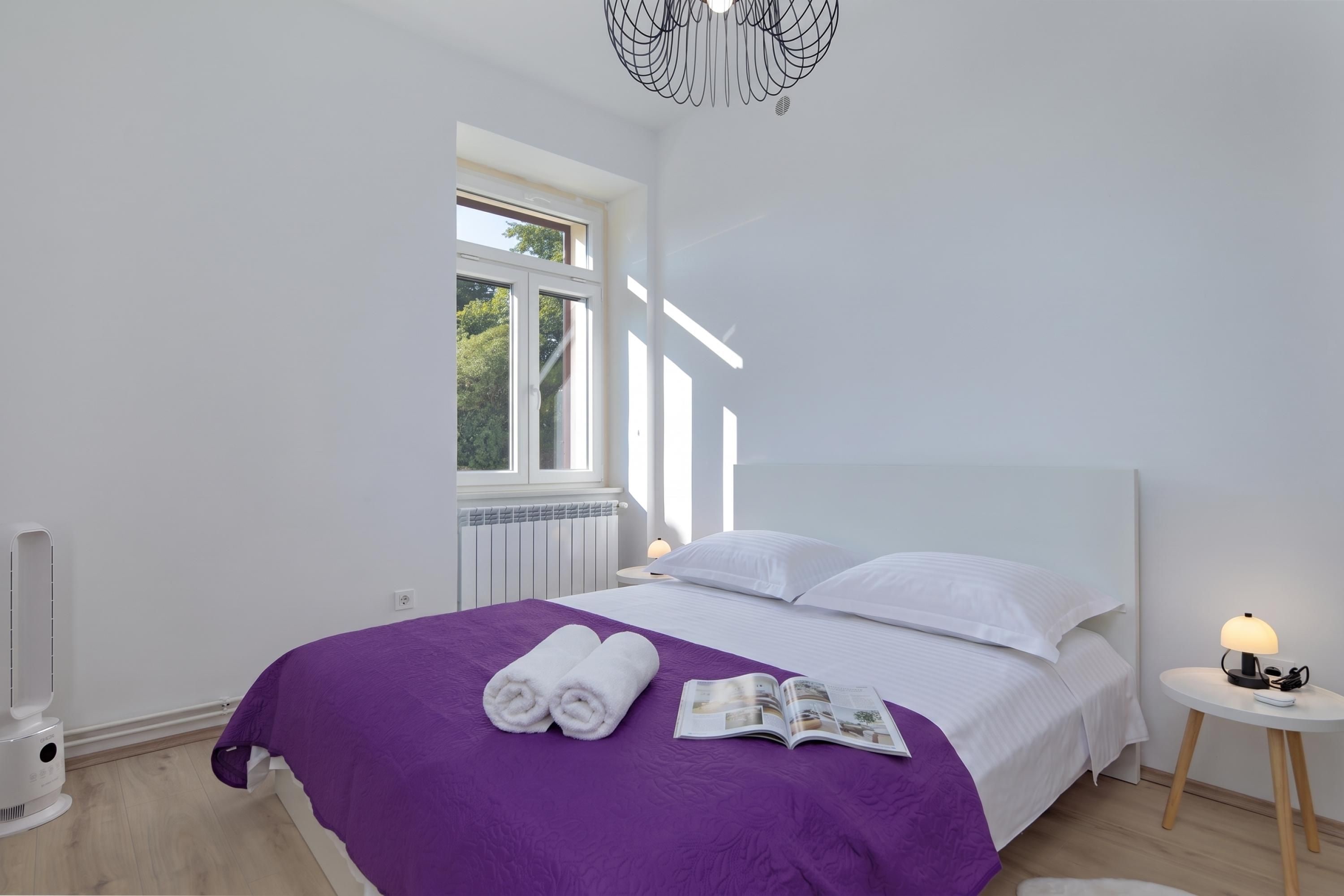 A bedroom with a large bed, a window, a radiator, a heater, and a bedside table with a lamp.