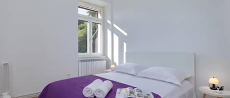 A bedroom with a large bed, a window, a radiator, a heater, and a bedside table with a lamp.