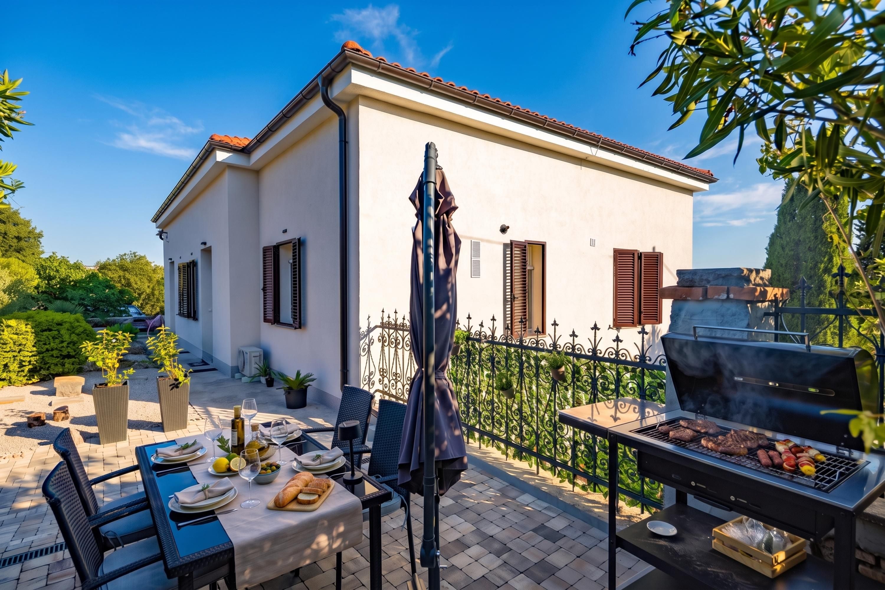 A patio with a dining table set for two, a grill, and a house with a white facade and brown shutters.