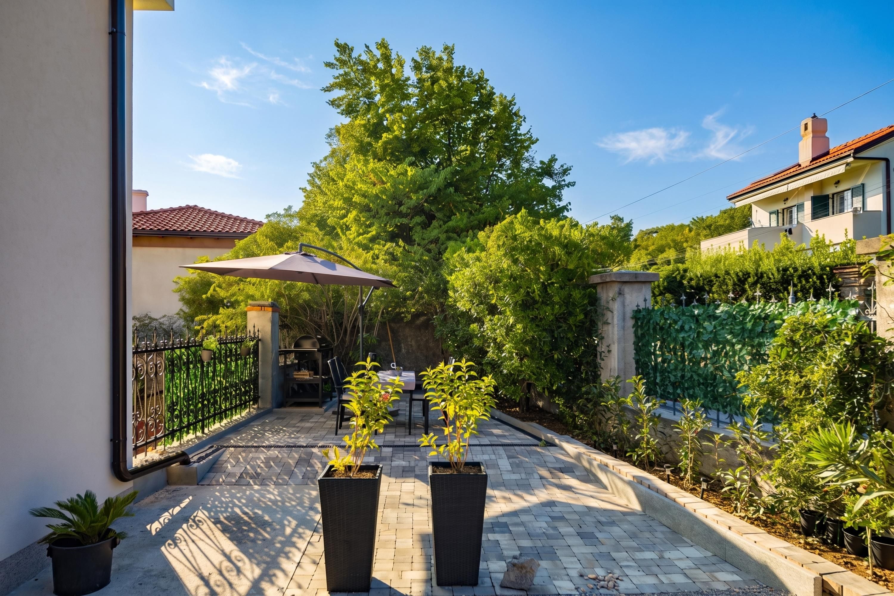 A paved courtyard with a table and chairs, surrounded by greenery and a building with a tiled roof.