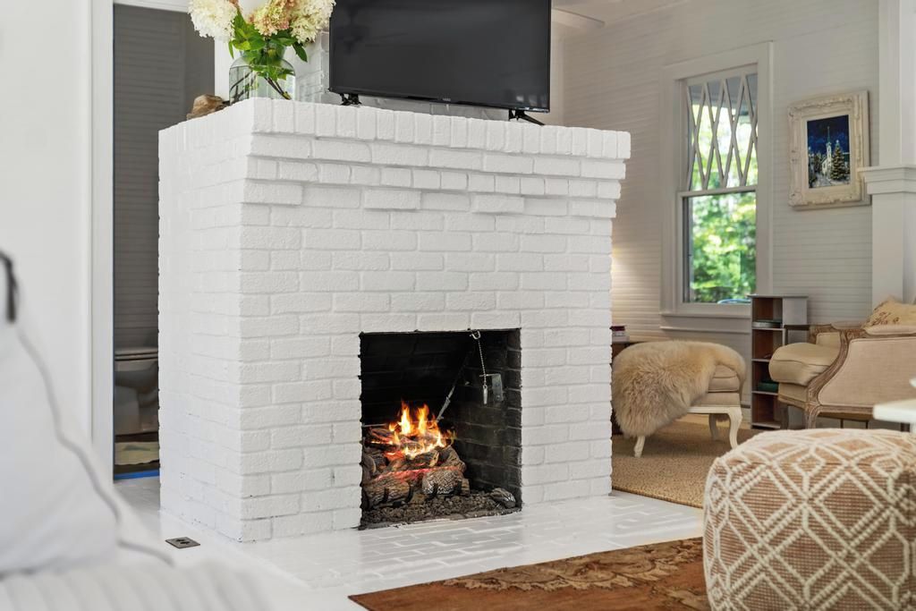 A white brick fireplace with a TV mounted above it, a fluffy white dog nearby, and a window with a view of greenery.