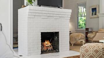 A white brick fireplace with a TV mounted above it, a fluffy white dog nearby, and a window with a view of greenery.