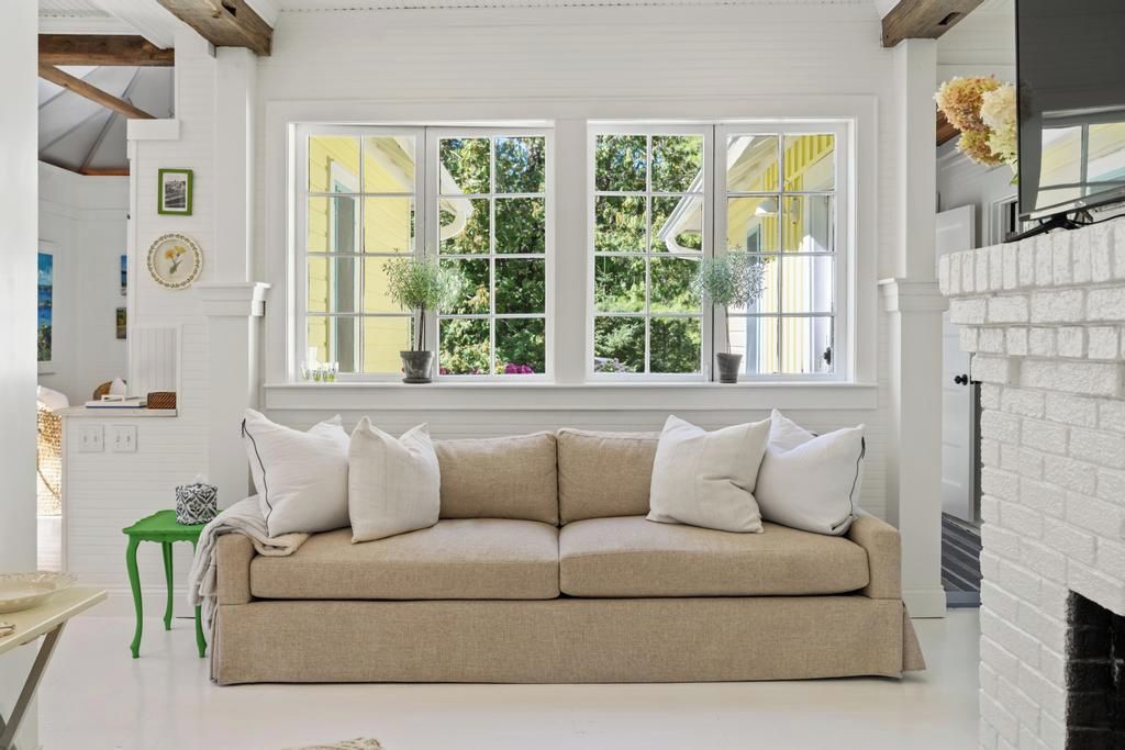 A beige sofa with white cushions in a room with a white fireplace, a green side table, and a window with yellow shutters.