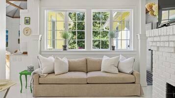 A beige sofa with white cushions in a room with a white fireplace, a green side table, and a window with yellow shutters.