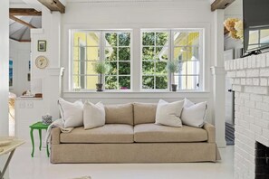 Een beige bank met witte kussens in een kamer met een witte schoorsteen, een groen bijzettafeltje en een raam met gele luiken.