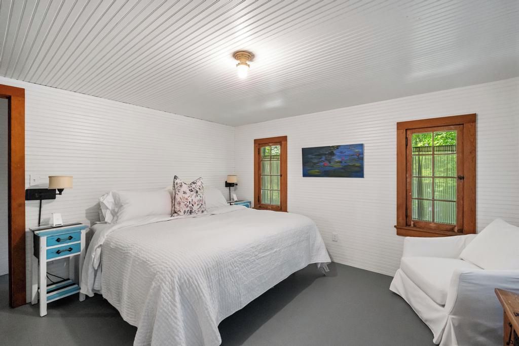 Un dormitorio con cama, mesitas de noche, una silla y dos ventanas con contraventanas.