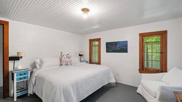 Un dormitorio con cama, mesitas de noche, una silla y dos ventanas con contraventanas.