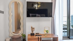 A modern living room with a large mirror, a flat-screen TV mounted on the wall, a wooden coffee table with decorative items, and a view of the city through large windows. - StayShort - Studio in Business Bay Sleeps 2 with Burj Views! (Dubai)