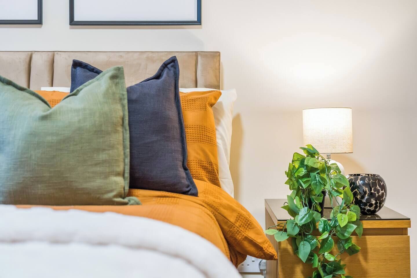 A neatly made bed with multiple pillows, a bedside lamp, and a small plant on a wooden nightstand.