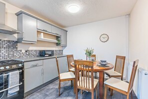 A compact kitchen with a stove, microwave, and cabinets, a dining table with chairs, and a wall clock. - #109 Modern 3-Bed Home in Moseley (Balsall Heath)