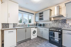 A compact kitchen with a dishwasher, washing machine, stove, and a window providing natural light. - #109 Modern 3-Bed Home in Moseley (Balsall Heath)