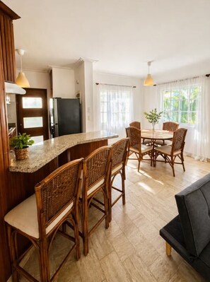 Una cocina con comedor, sillas de madera, un sofá negro y una planta en maceta.