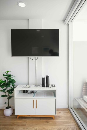 A modern living room with a mounted flat-screen TV, a white entertainment unit with drawers, a potted plant, and a glass door leading outside. - Just Renovated Marine Surf Corner Unit with Free Parking! (Honolulu)