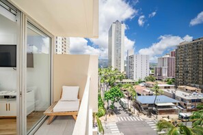 A balcony with a white cushioned chair and a glass door leading to a room with a bed and a desk. - Just Renovated Marine Surf Corner Unit with Free Parking! (Honolulu)